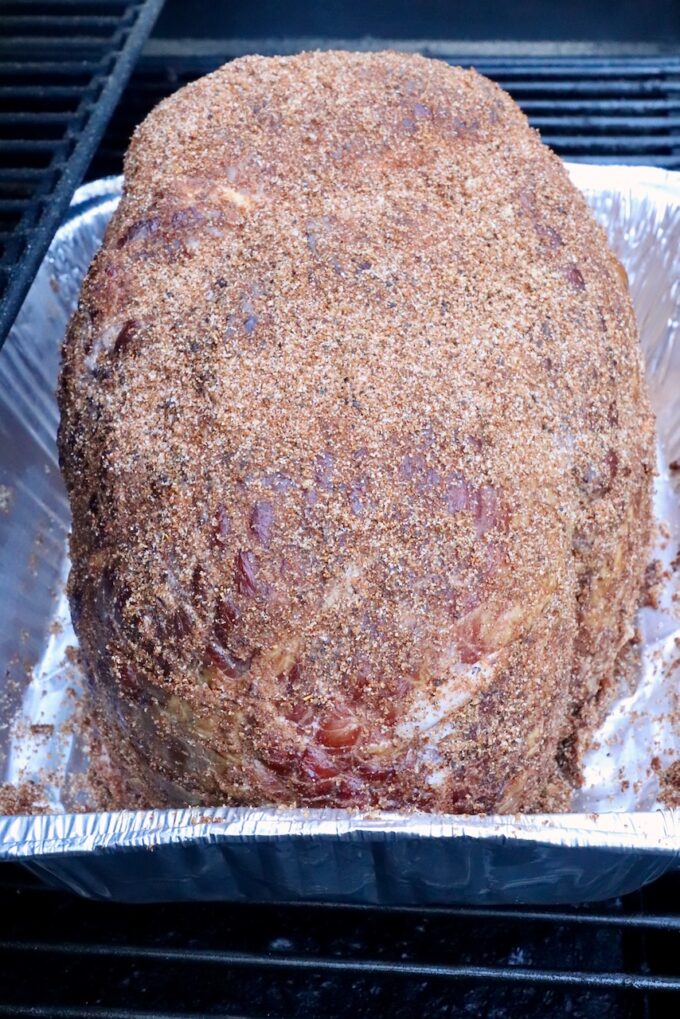 A seasoned ham covered in a brown sugar spice rub rests in a foil tray on a smoker. 