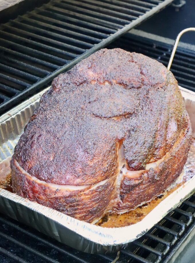 A large, well-seasoned ham with a crispy, dark crust sits in a foil pan on a smoker.