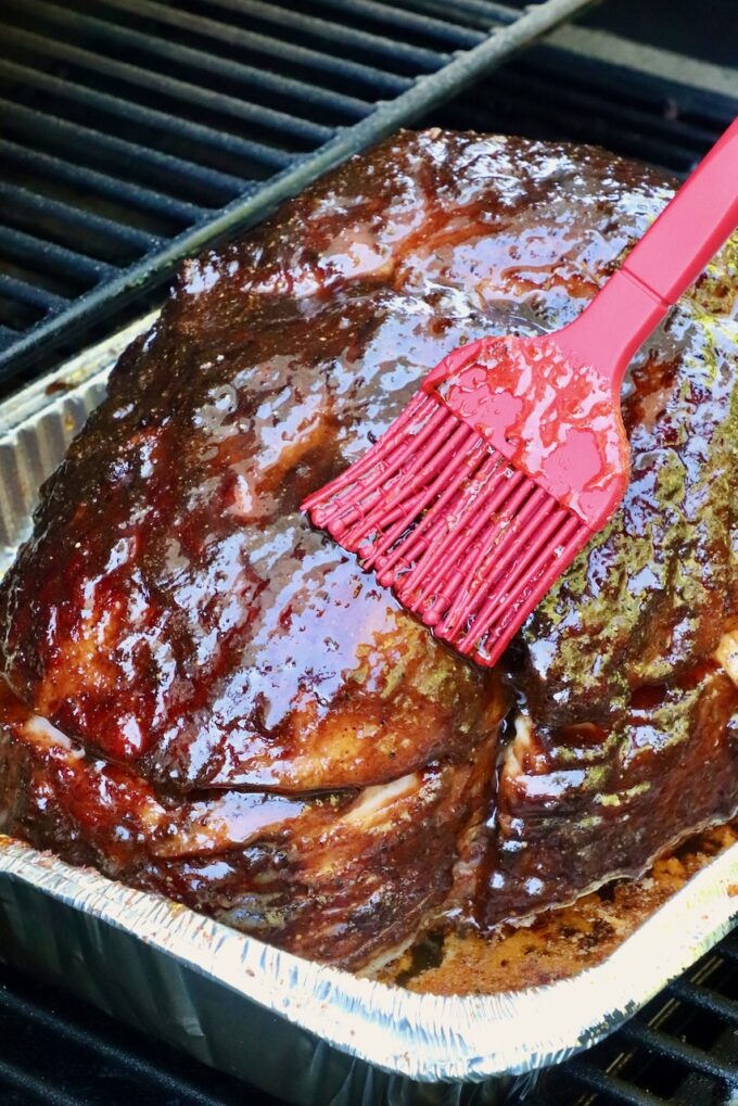 A smoked ham in a foil tray is being glazed with sauce using a red brush, on a smoker. 