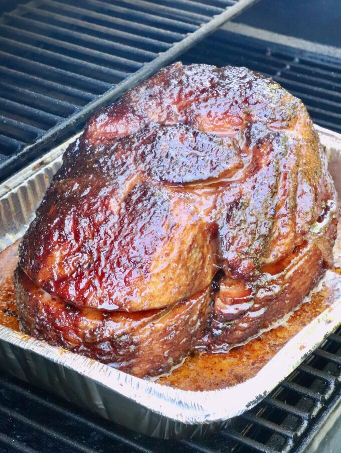 smoked ham with a glossy, caramelized glaze sits in a foil tray on a smoker