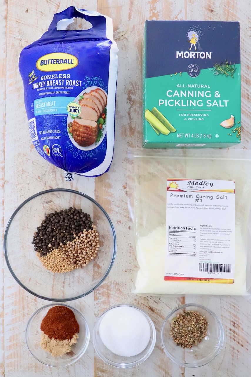 Ingredients for turkey pastrami include a package of turkey breast, canning salt, curing salt, and bowls of spices like peppercorns, mustard seeds, and paprika.