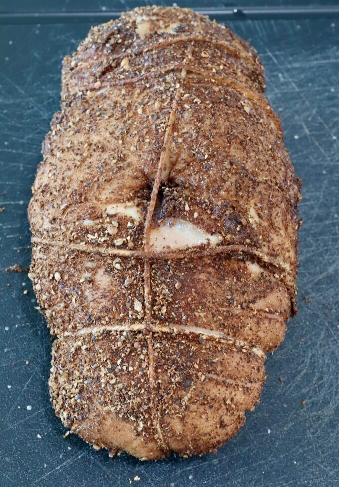 turkey breast tied with twine, coated in a dark, textured spice rub, lies on a black cutting board.