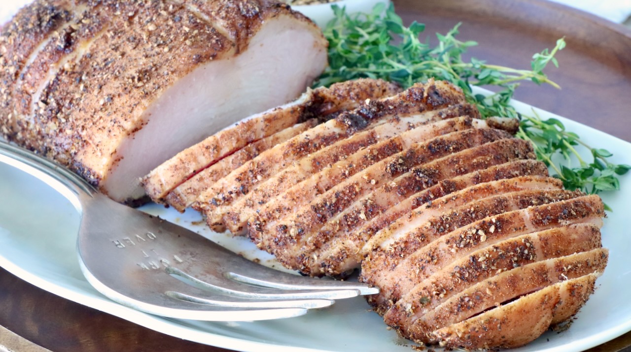 Sliced smoked turkey breast seasoned with spices on a white plate, garnished with fresh thyme. A silver carving fork rests beside it.