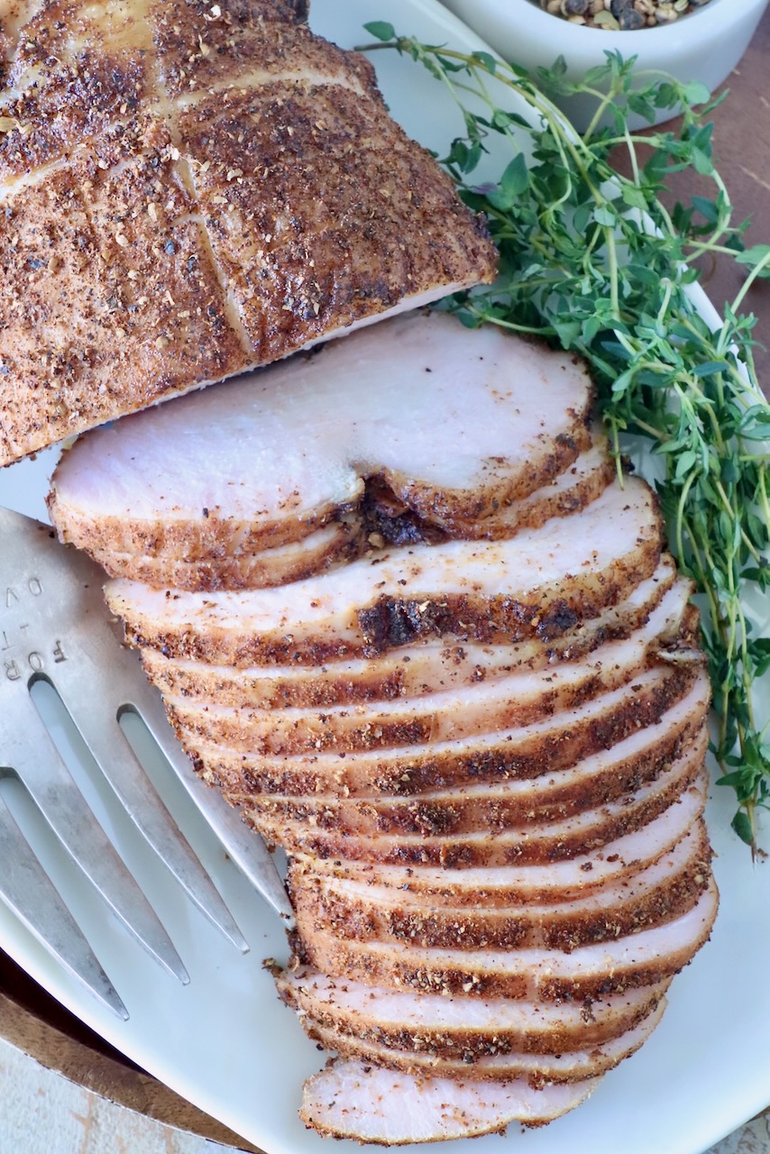 Sliced smoked turkey breast on a white platter, seasoned with spices. Fresh herbs are placed beside it, along with a serving fork.
