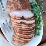 Sliced smoked turkey breast on a white platter, seasoned with spices. Fresh herbs are placed beside it, along with a serving fork.