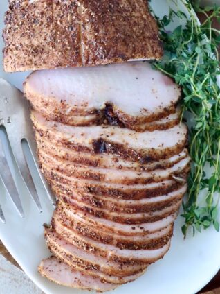 Sliced smoked turkey breast on a white platter, seasoned with spices. Fresh herbs are placed beside it, along with a serving fork.