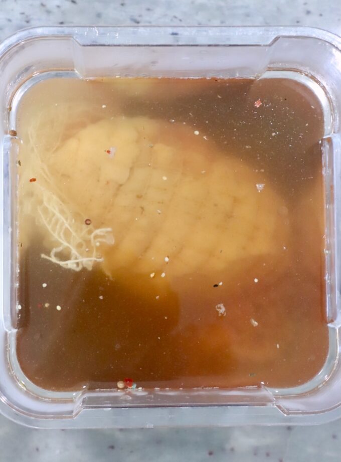 A turkey breast is submerged in a light brown brine within a plastic container.