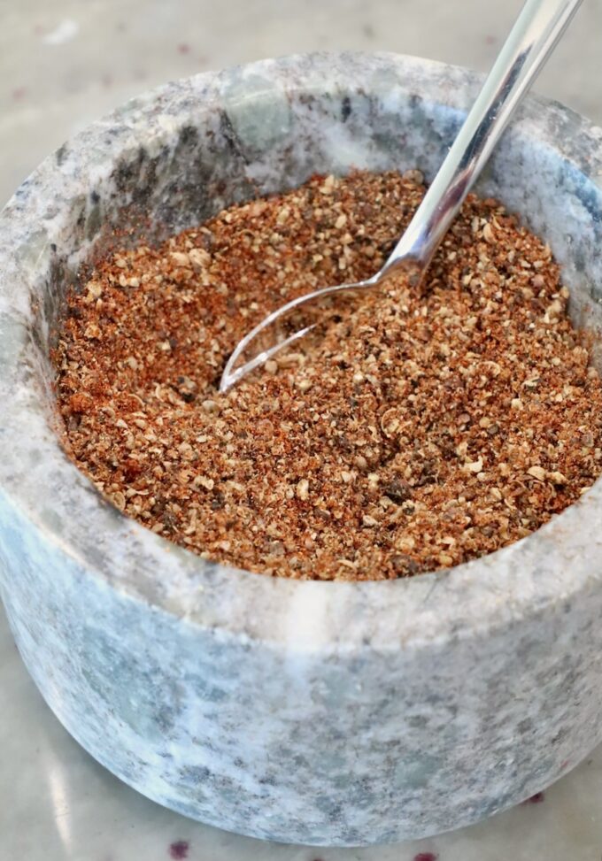 A close-up of a marble mortar filled with a coarse, mixed spice blend. A shiny metal spoon is partially submerged in the spices