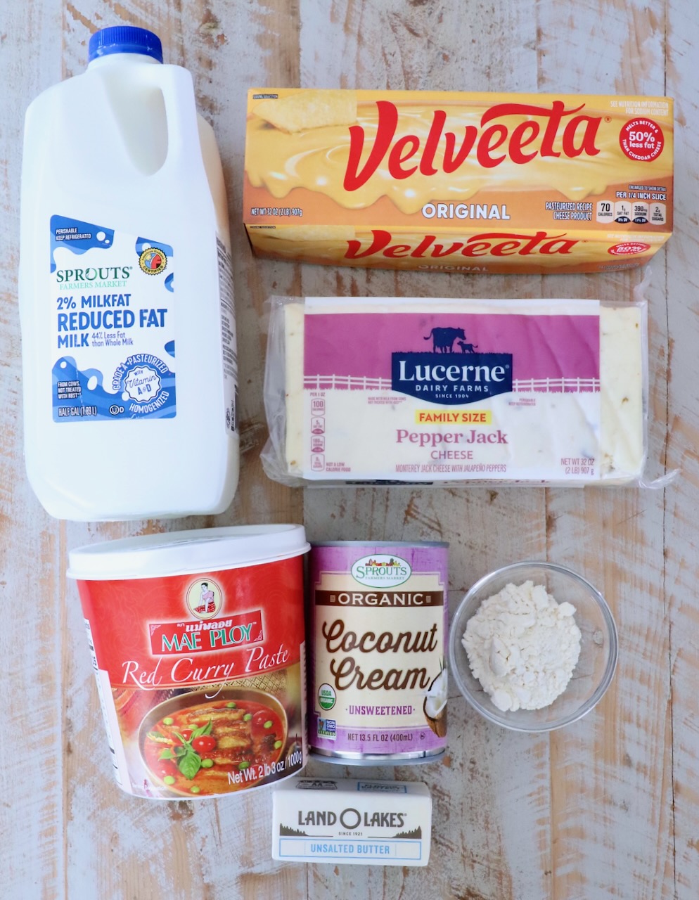 A flat lay of cooking ingredients on a wooden surface. Items include milk, Velveeta cheese, pepper jack cheese, red curry paste, coconut cream, flour, and a stick of butter.