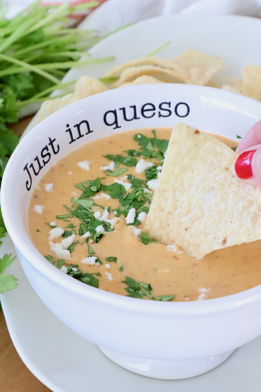 A bowl of queso dip is garnished with cilantro and cheese, labeled "Just in queso." A hand with red nail polish dips a tortilla chip in the dip. Fresh cilantro and chips are in the background.