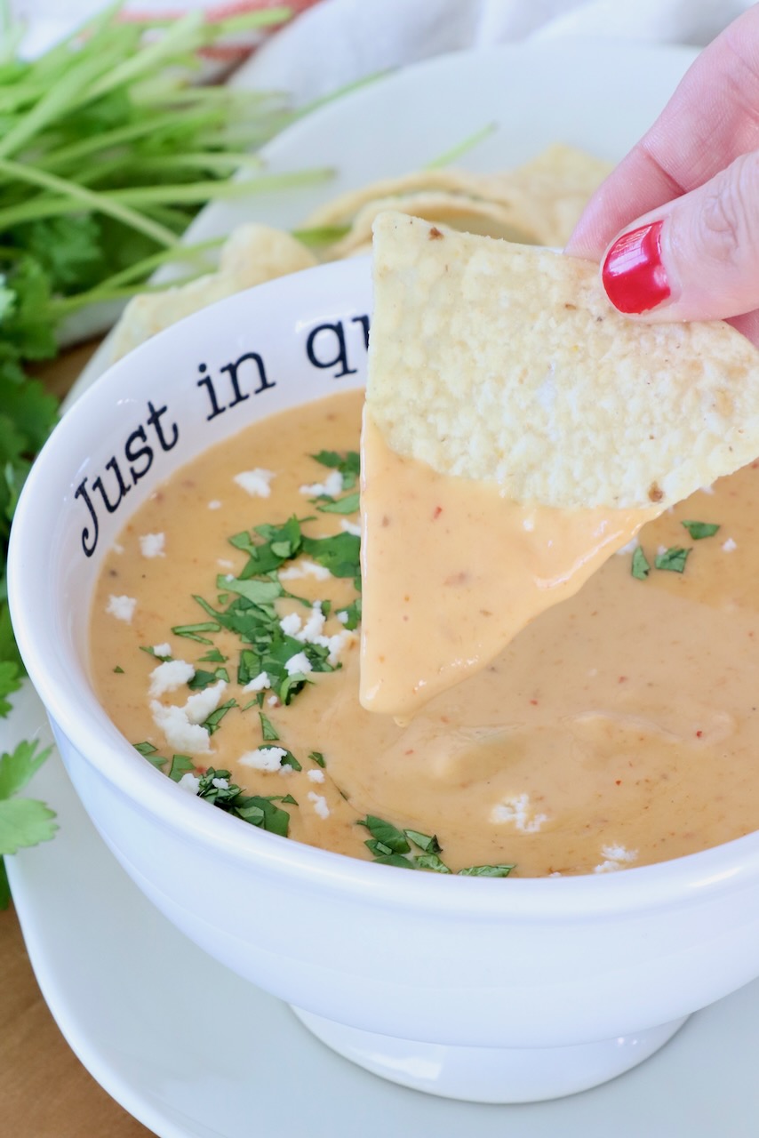 A hand with red nail polish dips a tortilla chip into creamy queso garnished with cilantro and queso fresco. A bowl reads "Just in queso."