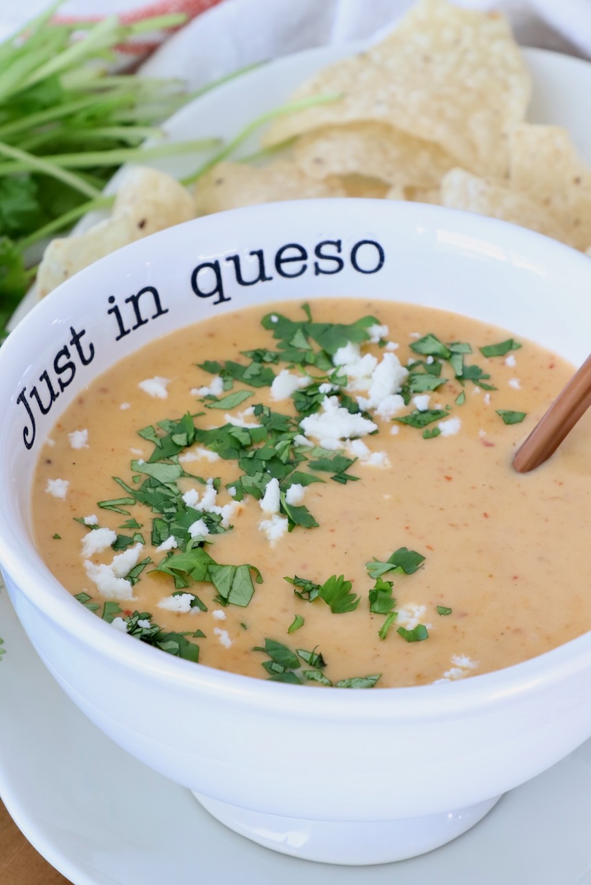 A bowl of creamy queso topped with chopped cilantro and crumbled cheese. The bowl has "Just in queso" written on it and is paired with tortilla chips.
