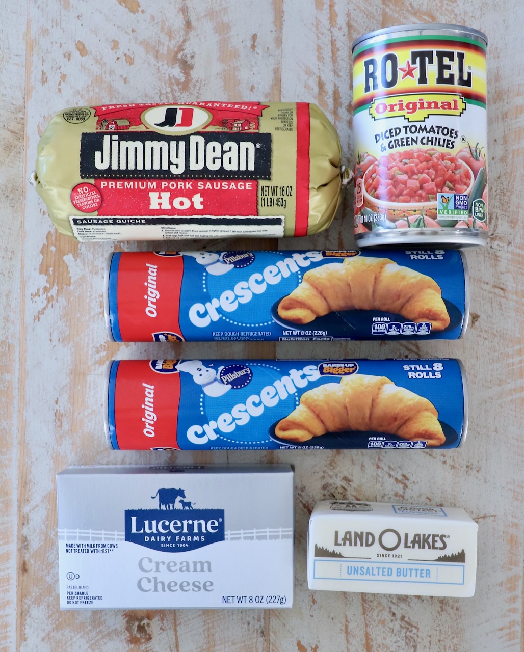 Assorted grocery items on a wooden surface: Jimmy Dean pork sausage, Rotel tomatoes, two Pillsbury crescent roll cans, Lucerne cream cheese, Land O'Lakes butter.