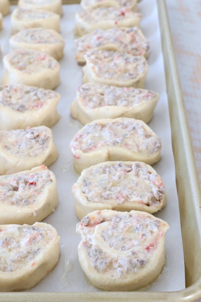 Rows of uncooked pastry pinwheels filled with a creamy mixture of sausage and tomatoes are neatly arranged on a parchment-lined baking sheet.