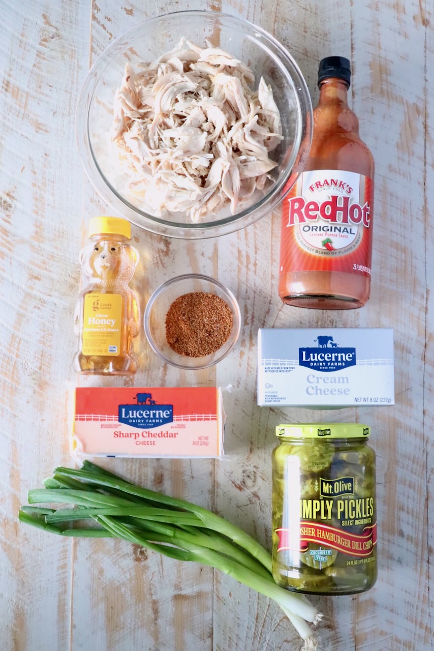 A wooden surface displays ingredients including: shredded chicken, hot sauce, pickles, green onions, honey, cream cheese, cheddar, and seasoning.