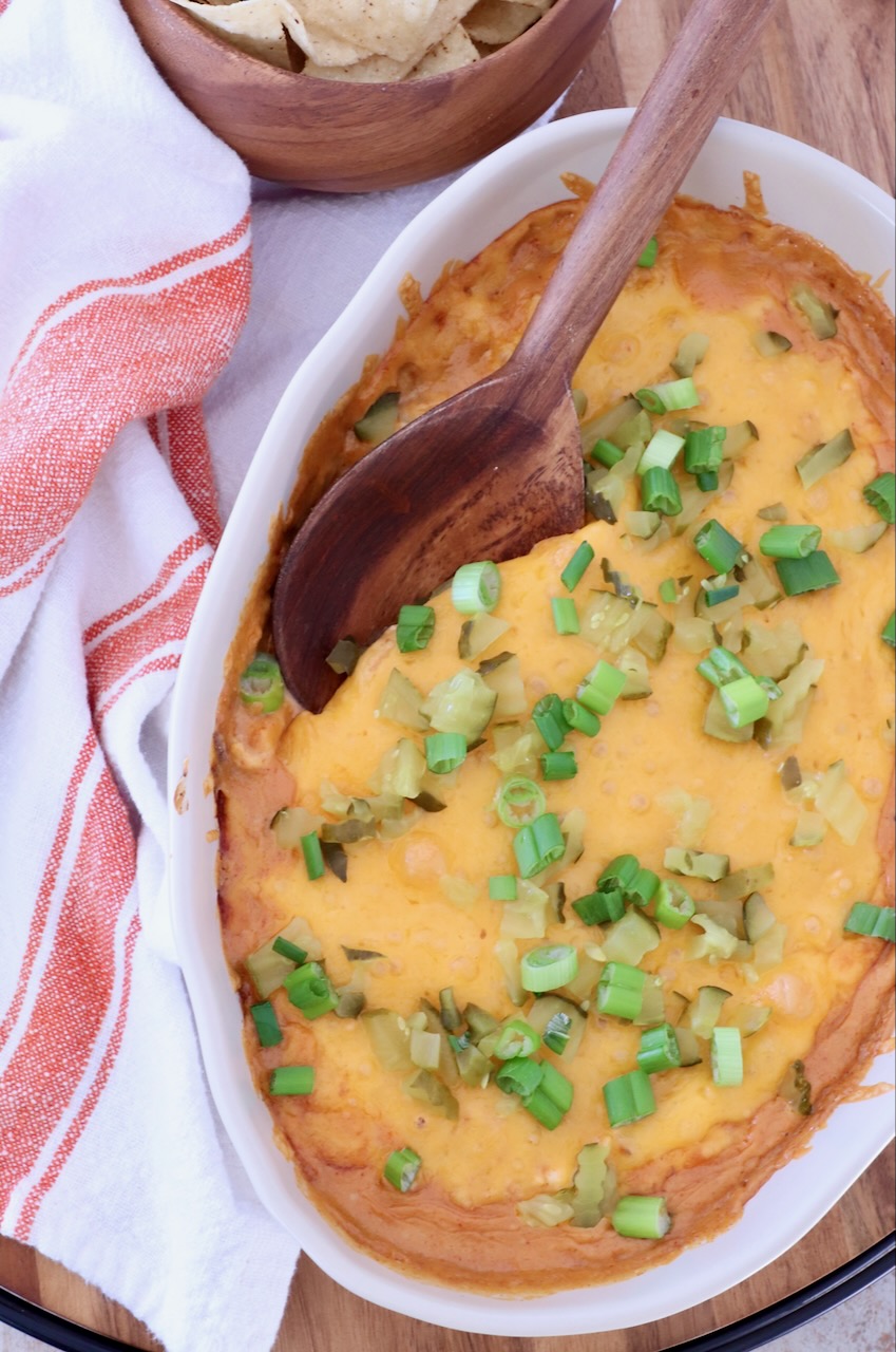 Close-up of a baked cheesy dip in an oval dish topped with green onions and pickles. A wooden spoon rests on top. Tortilla chips in a bowl and a striped towel are nearby.