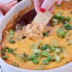 A hand with red nails dips a tortilla chip into a cheesy, baked dip topped with chopped green onions, in a white dish
