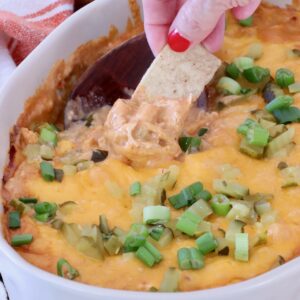 A hand with red nails dips a tortilla chip into a cheesy, baked dip topped with chopped green onions, in a white dish