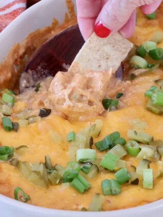 A hand with red nails dips a tortilla chip into a cheesy, baked dip topped with chopped green onions, in a white dish