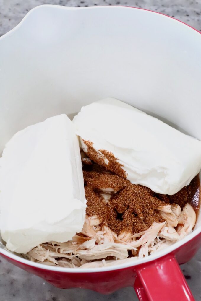 A red mixing bowl with shredded chicken, two blocks of cream cheese, and a sprinkle of spices.