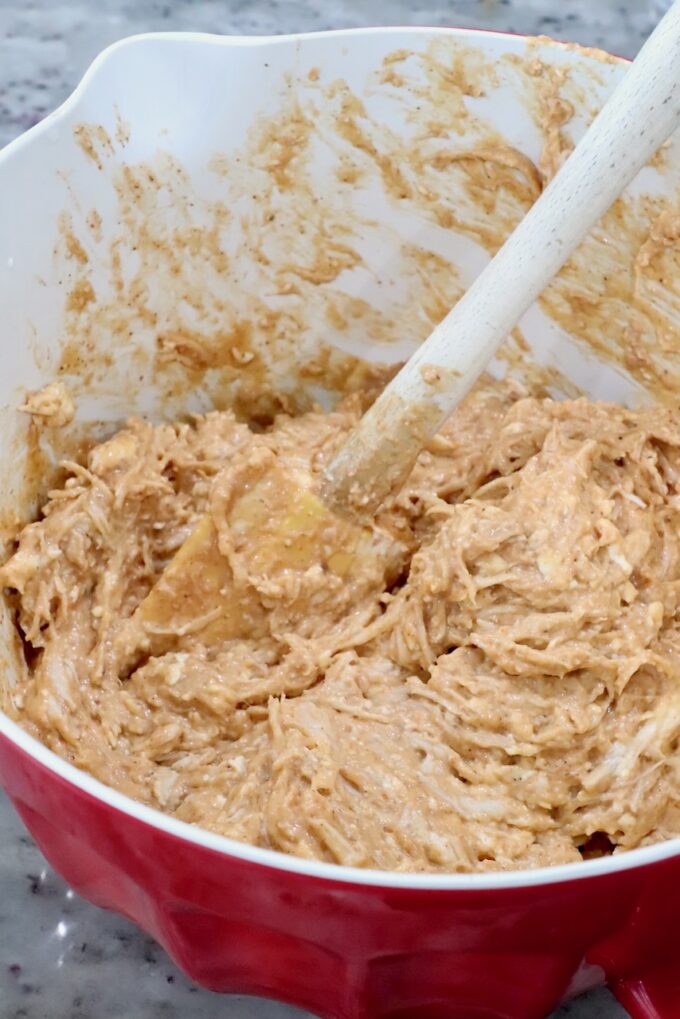 A red bowl filled with a thick, creamy chicken and cheese mixture being mixed with a wooden spoon.