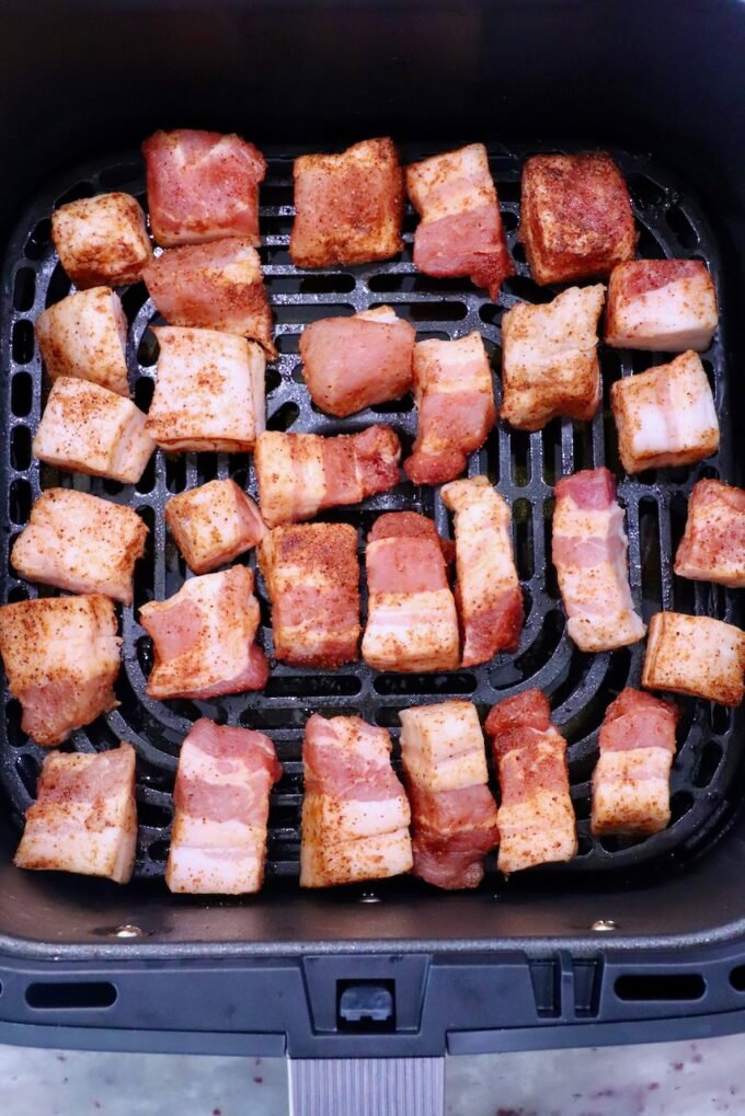 Cubed pork belly pieces, seasoned with spices, are evenly spread in the basket of an air fryer. The meat is pink and golden, ready for cooking.