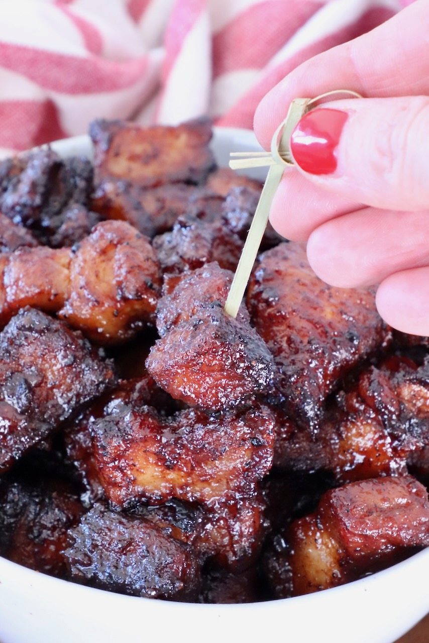 A hand with red nail polish holds a toothpick, picking up a glazed, caramelized pork belly bite from a bowl filled with similar pieces. Red-striped cloth is in the background.