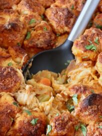 Close-up of a buffalo chicken pot pie with a serving spoon. The pie has golden-brown biscuit topping and is garnished with herbs.