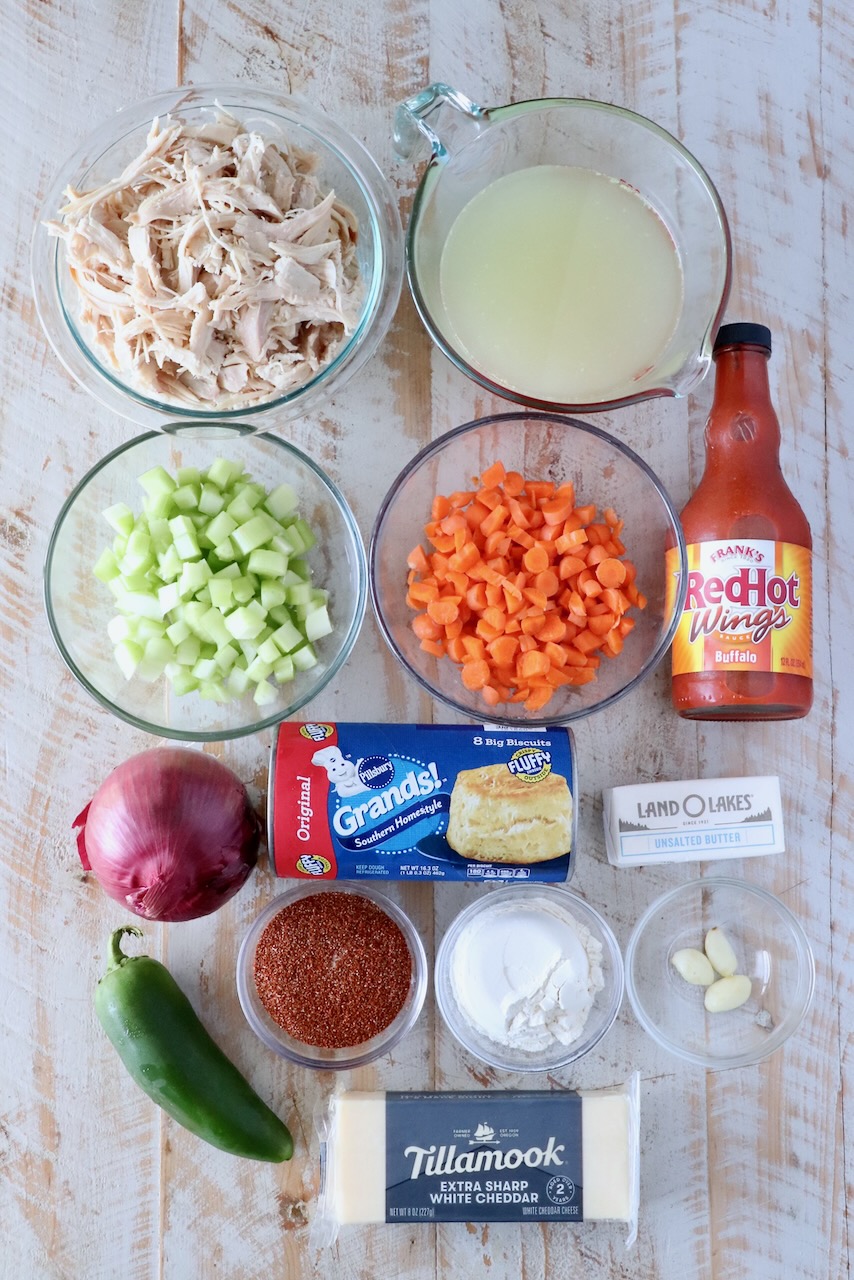 Overhead view of cooking ingredients: shredded chicken, broth, diced celery, carrots, hot sauce, onion, jalapeño, biscuits, butter, seasonings, and cheddar cheese