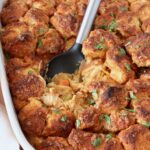 Close-up of a baked buffalo chicken pot pie topped with golden biscuit pieces. Garnished with fresh herbs, a serving spoon rests in the dish.