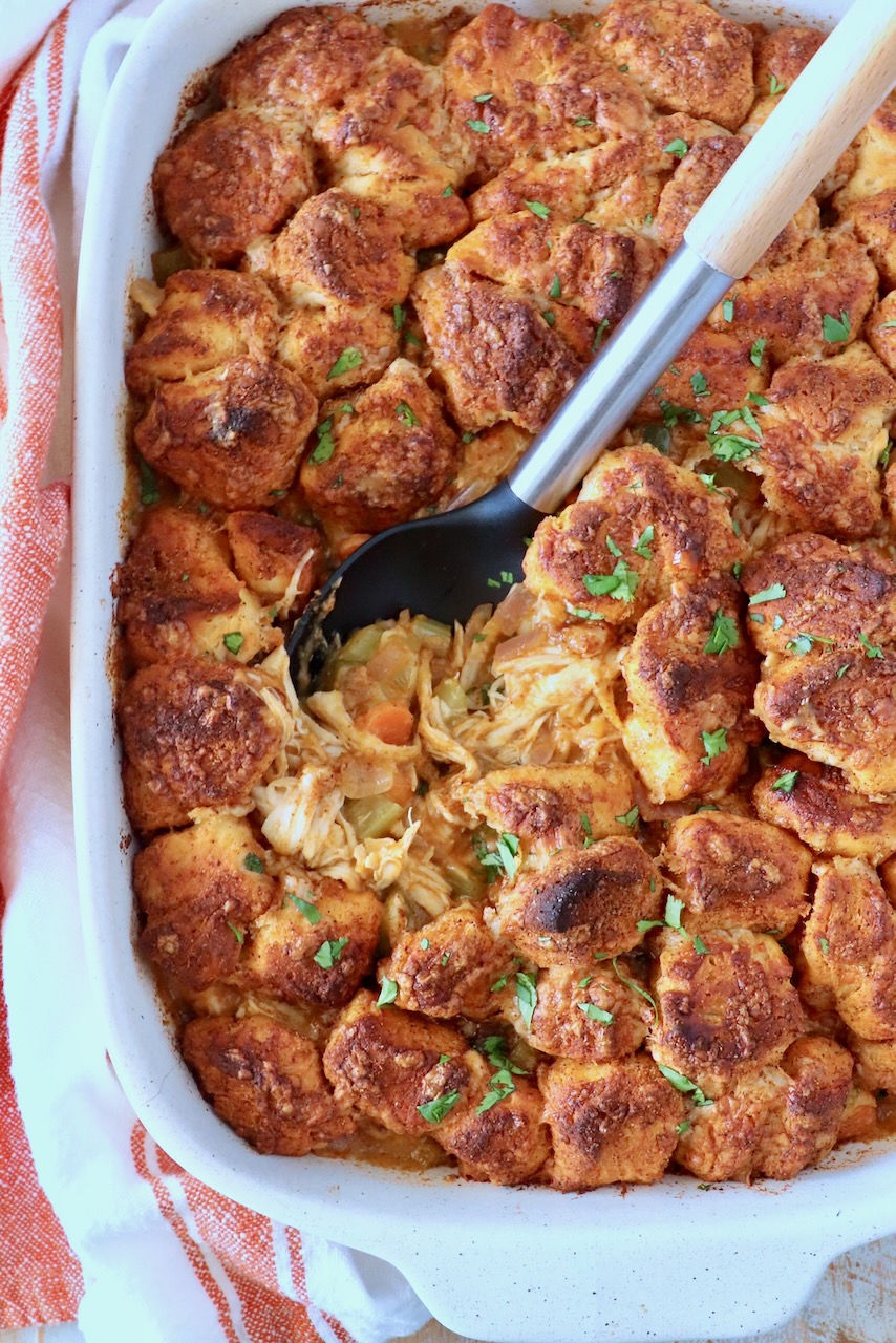 Golden-brown biscuits topped with herbs cover a hearty chicken and vegetable pot pie in a white dish. A spoon reveals the warm, savory filling.