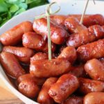 A close-up of glazed cocktail sausages in a white bowl with toothpicks, placed on a wooden table. Fresh cilantro is visible in the background.