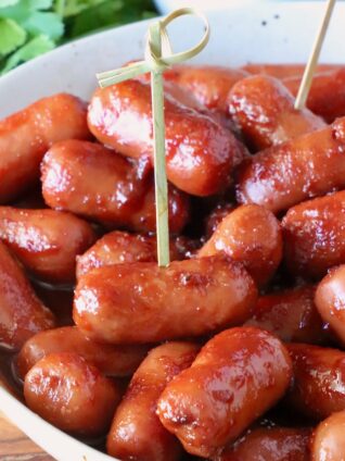 A close-up of glazed cocktail sausages in a white bowl with toothpicks, placed on a wooden table. Fresh cilantro is visible in the background.