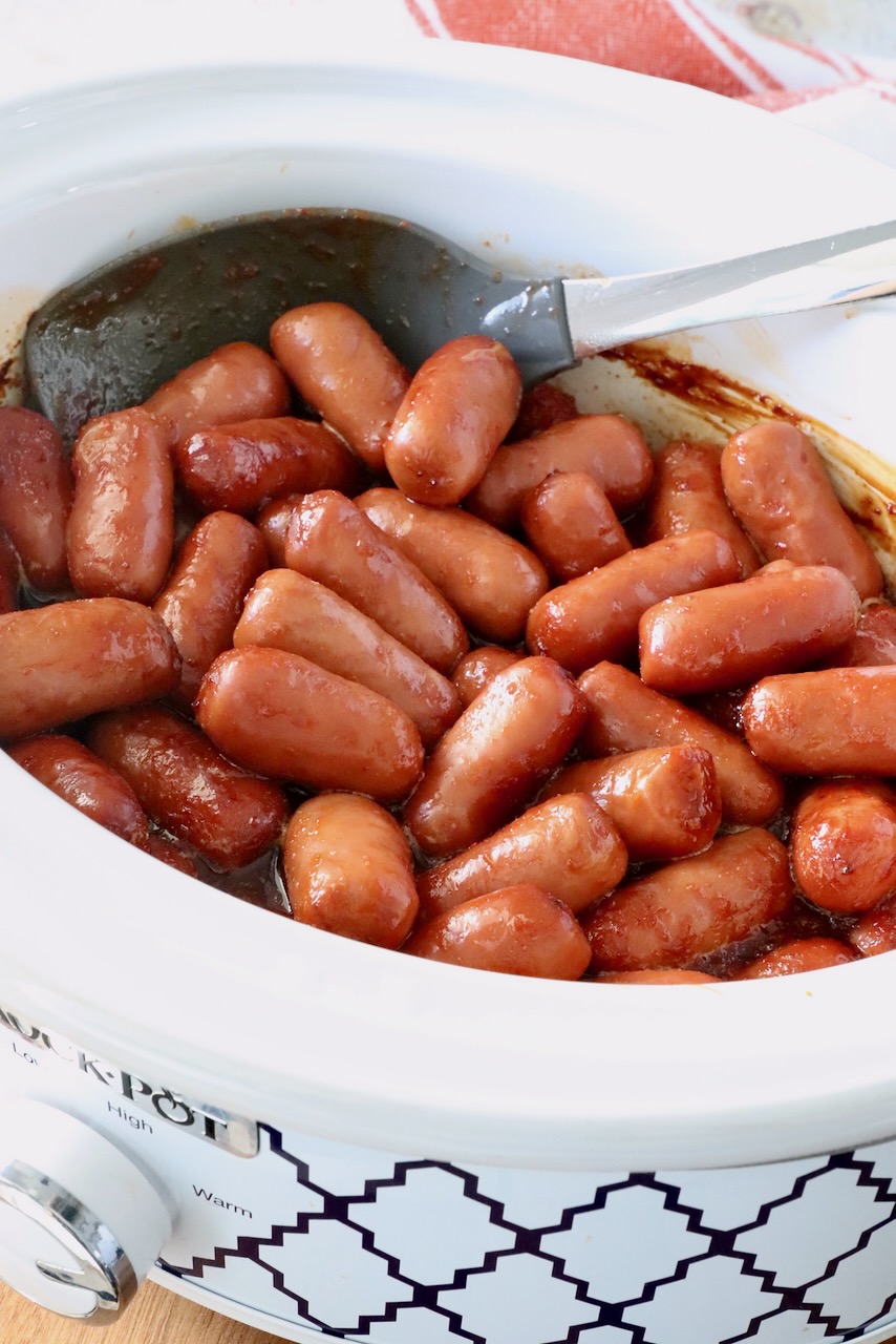 Slow cooker filled with glossy cocktail sausages in a chipotle sauce, with a serving spoon resting inside.