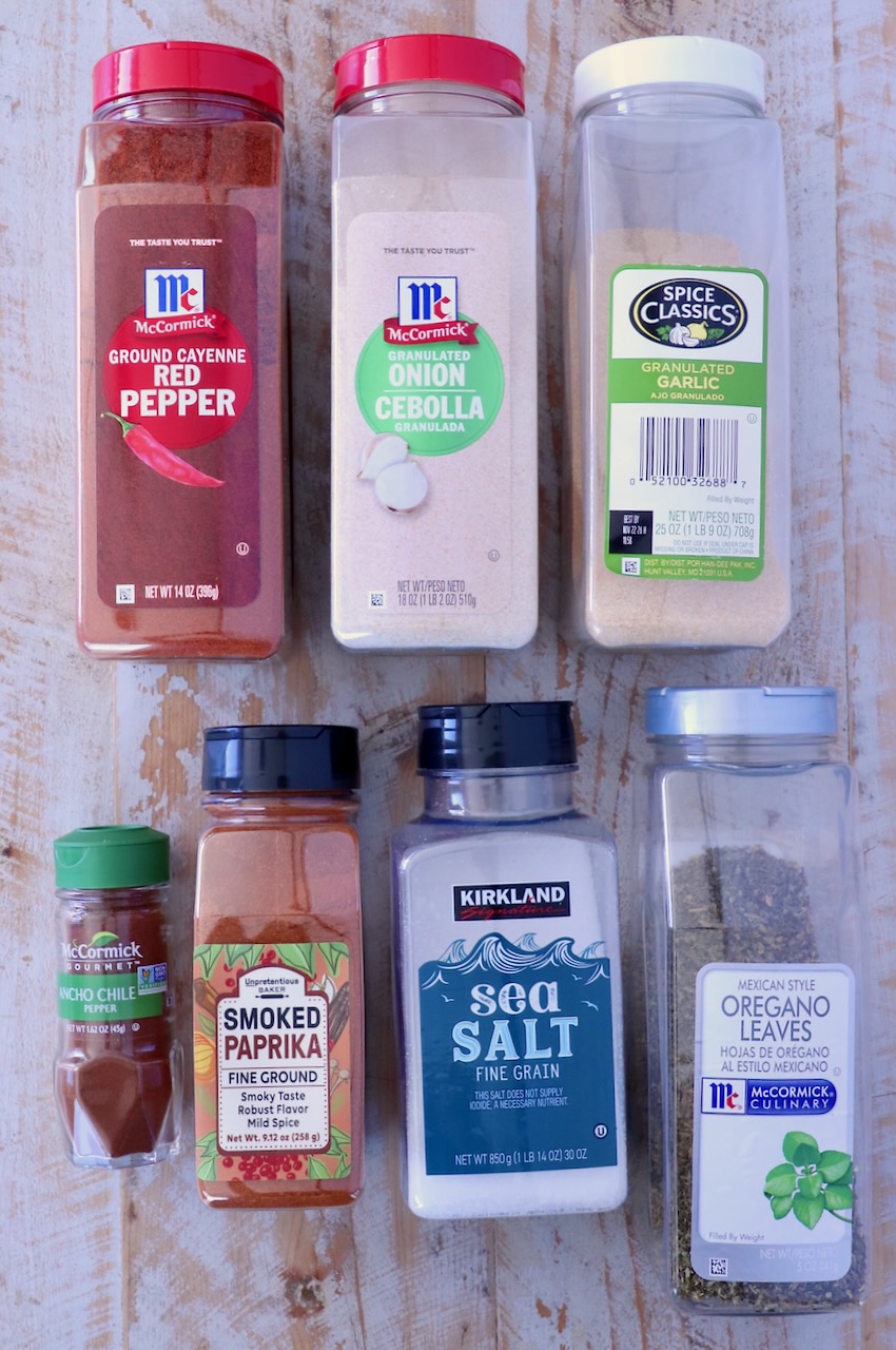 Assorted spice containers, including red pepper, onion powder, garlic, ancho chile, paprika, sea salt, and oregano, arranged on a wooden surface.