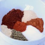 A white bowl containing vibrant heaps of spices, including red chili powder, paprika, oregano, and garlic powder, arranged in a colorful circle.