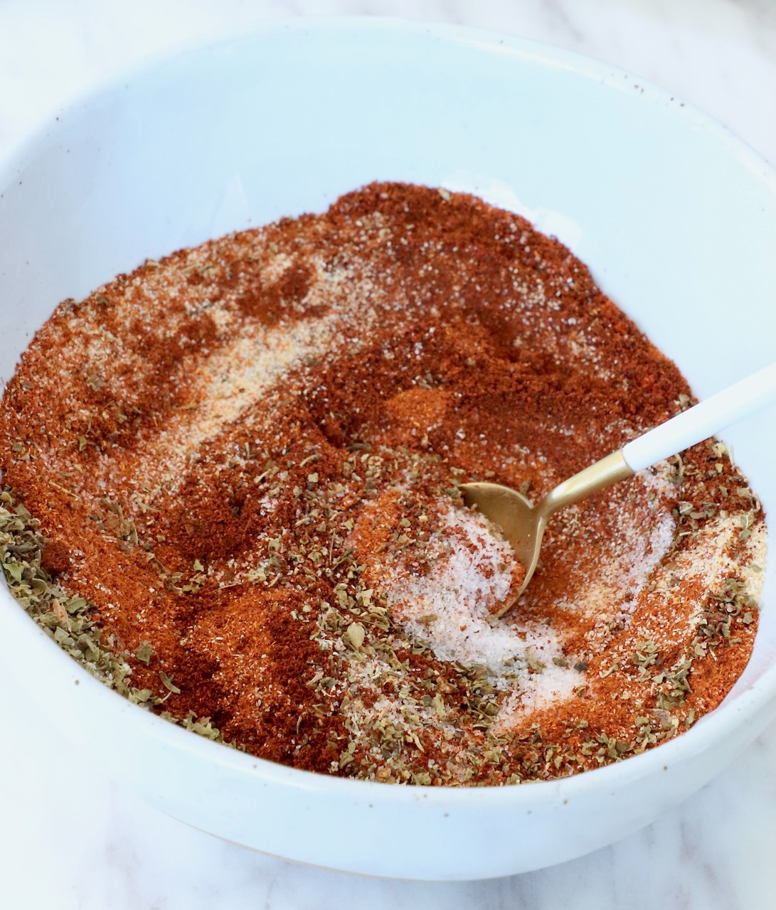 A white bowl filled with mixed spices including red paprika, herbs, and salt. A white-handled gold spoon rests in the mixture.