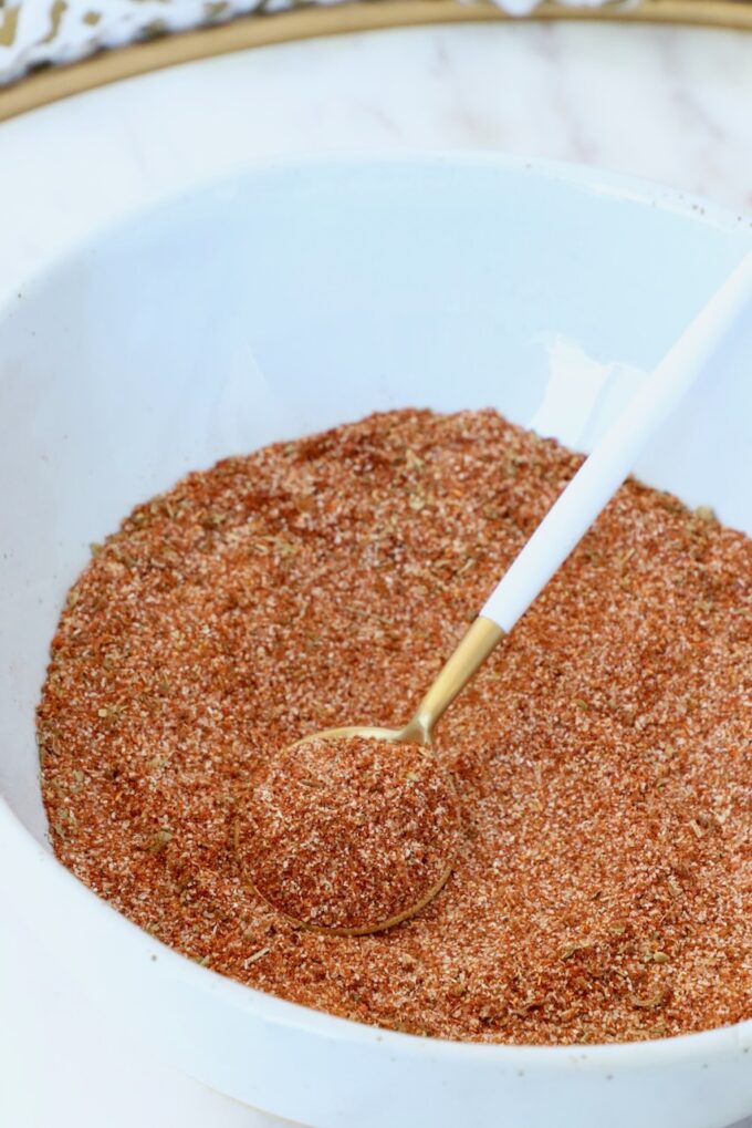 Close-up of a white bowl filled with reddish-brown spice mix. A gold and white spoon rests on top.