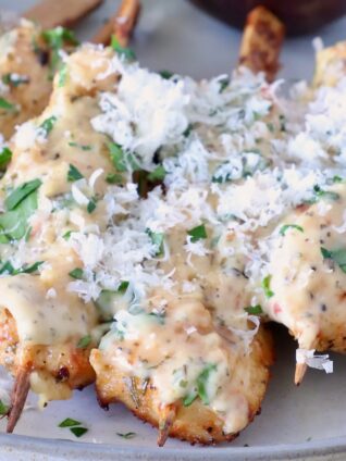 cooked chicken skewers drizzled with a creamy sauce, topped with shredded cheese and fresh herbs, on a light-colored plate.