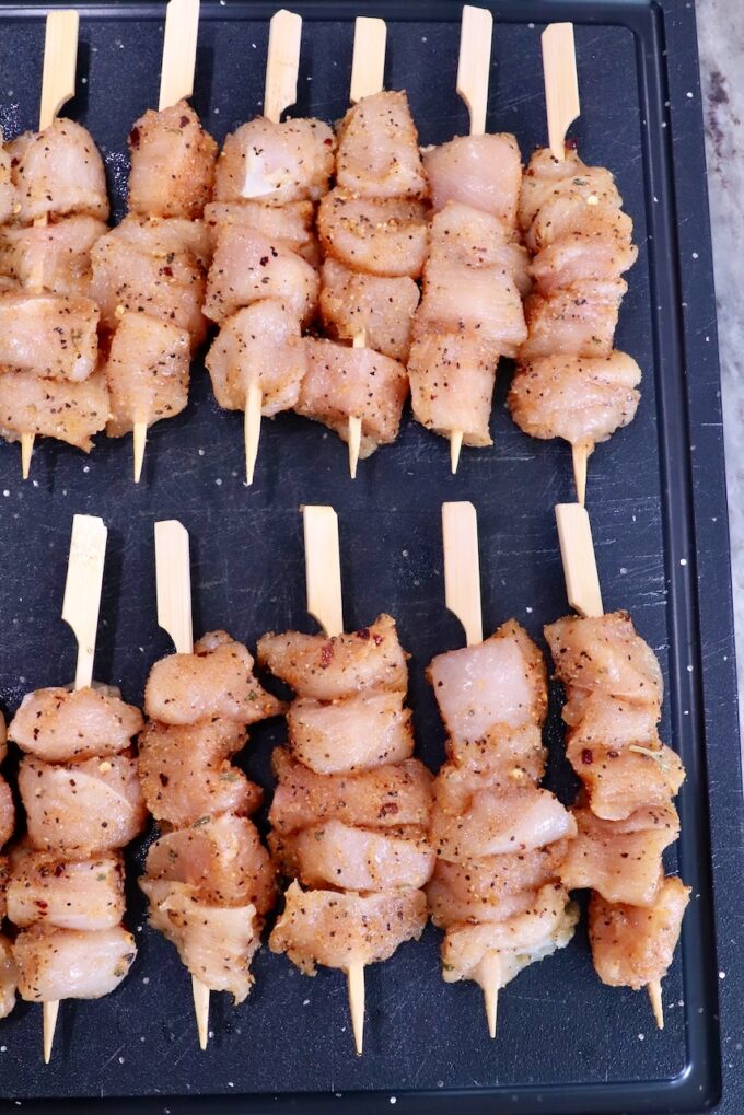 Raw chicken skewers seasoned with spices are neatly arranged on a black cutting board. The chicken is cut into chunks.