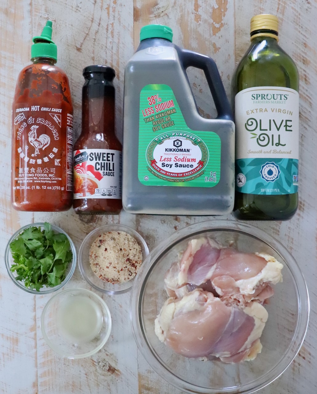 Ingredients for a recipe are arranged on a wooden surface. From left to right: Sriracha, sweet chili sauce, soy sauce, olive oil, cilantro, thai seasoning, lime juice, and raw chicken thighs.