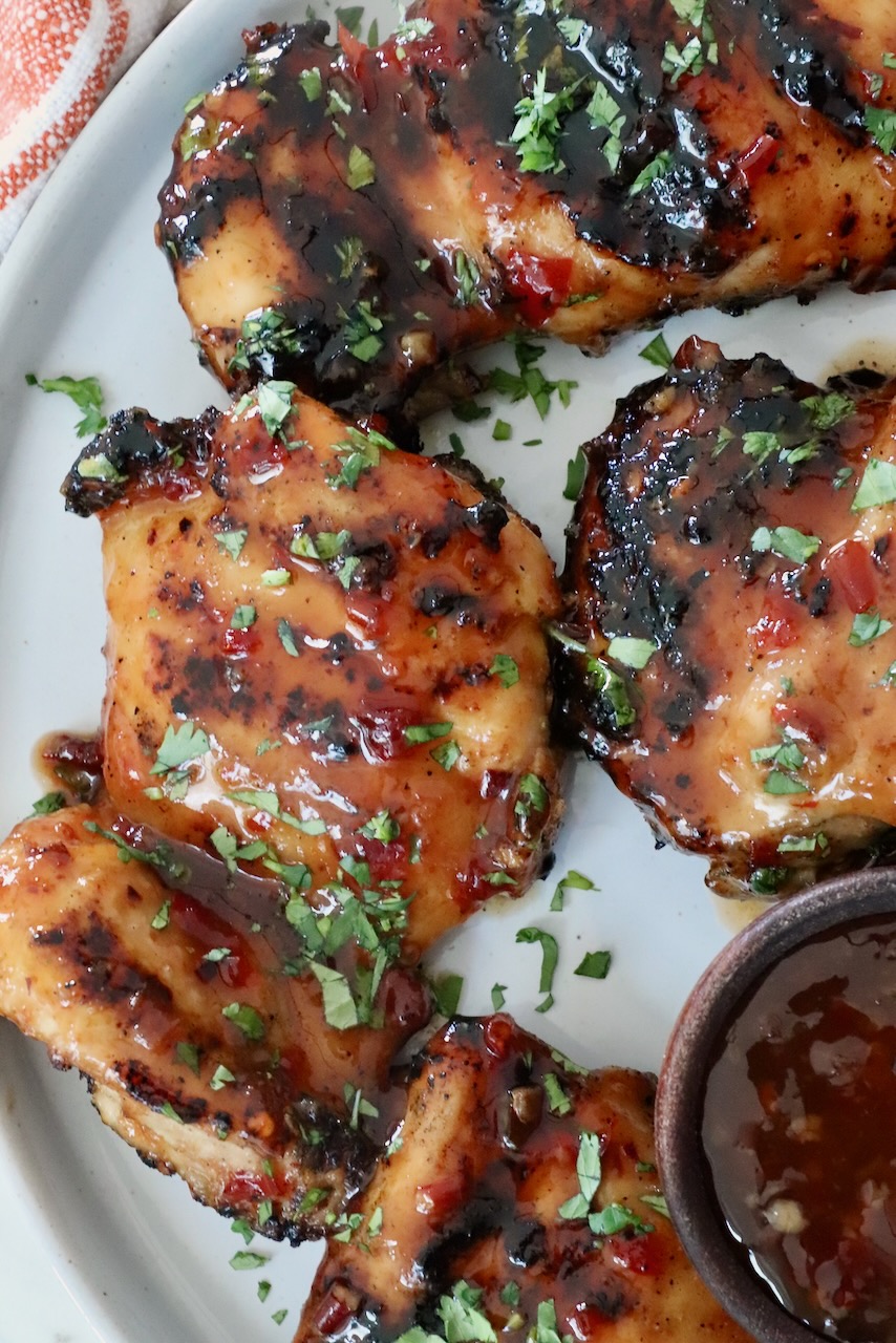 Grilled glazed chicken thighs are topped with fresh herbs on a white plate. A small bowl of dipping sauce is visible on the side.