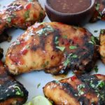 Grilled Thai glazed chicken thighs are topped with fresh herbs on a white plate. A small bowl of dipping sauce is visible on the side.