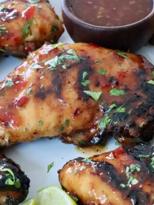 Grilled Thai glazed chicken thighs are topped with fresh herbs on a white plate. A small bowl of dipping sauce is visible on the side.