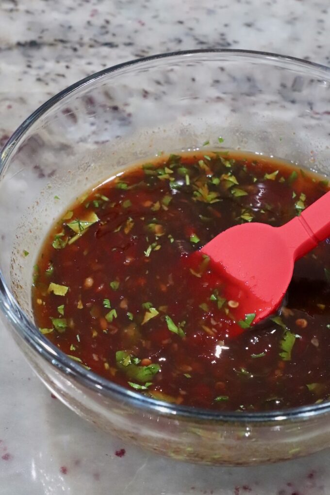 A glass bowl filled with a reddish-brown sauce, featuring chopped herbs, is on a marble surface. A red spatula rests in the bowl.
