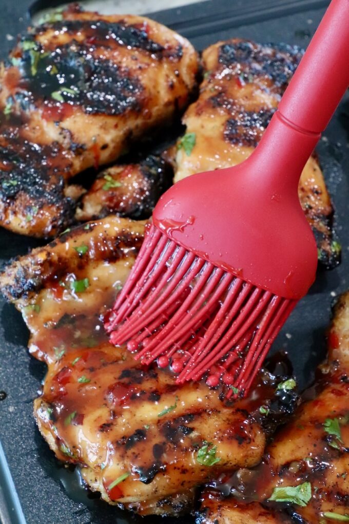 Grilled chicken thighs being brushed with glossy sauce using a red brush, showing charred marks and fresh herbs on a dark surface.