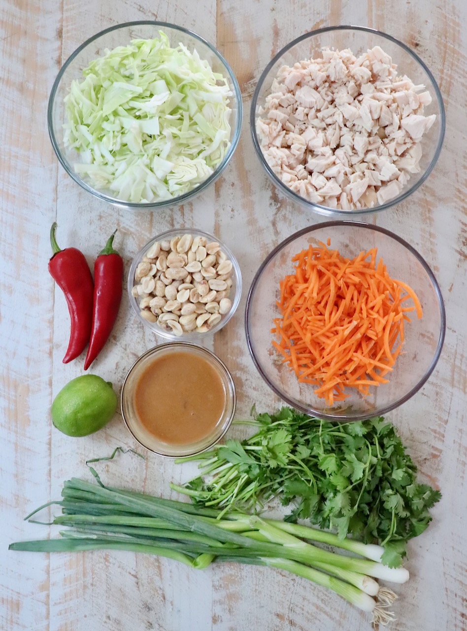 Bowls of shredded cabbage, diced chicken, and grated carrot on a wooden surface, surrounded by red chilies, peanuts, lime, cilantro, green onions, and peanut sauce.