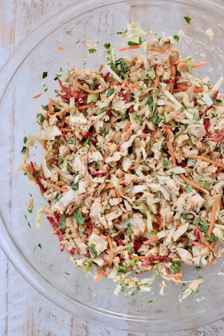A glass bowl filled with colorful chicken salad on a light wooden surface. The salad includes shredded cabbage, carrots, red peppers, and green herbs, giving a fresh, vibrant look.