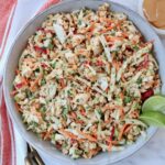 A vibrant Thai chicken salad in a bowl with shredded cabbage, carrots, peanuts, and cilantro. Two lime wedges are on top, and a creamy dressing is on the side.