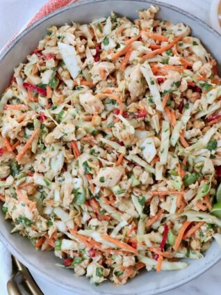 A vibrant Thai chicken salad in a bowl with shredded cabbage, carrots, peanuts, and cilantro. Two lime wedges are on top, and a creamy dressing is on the side.